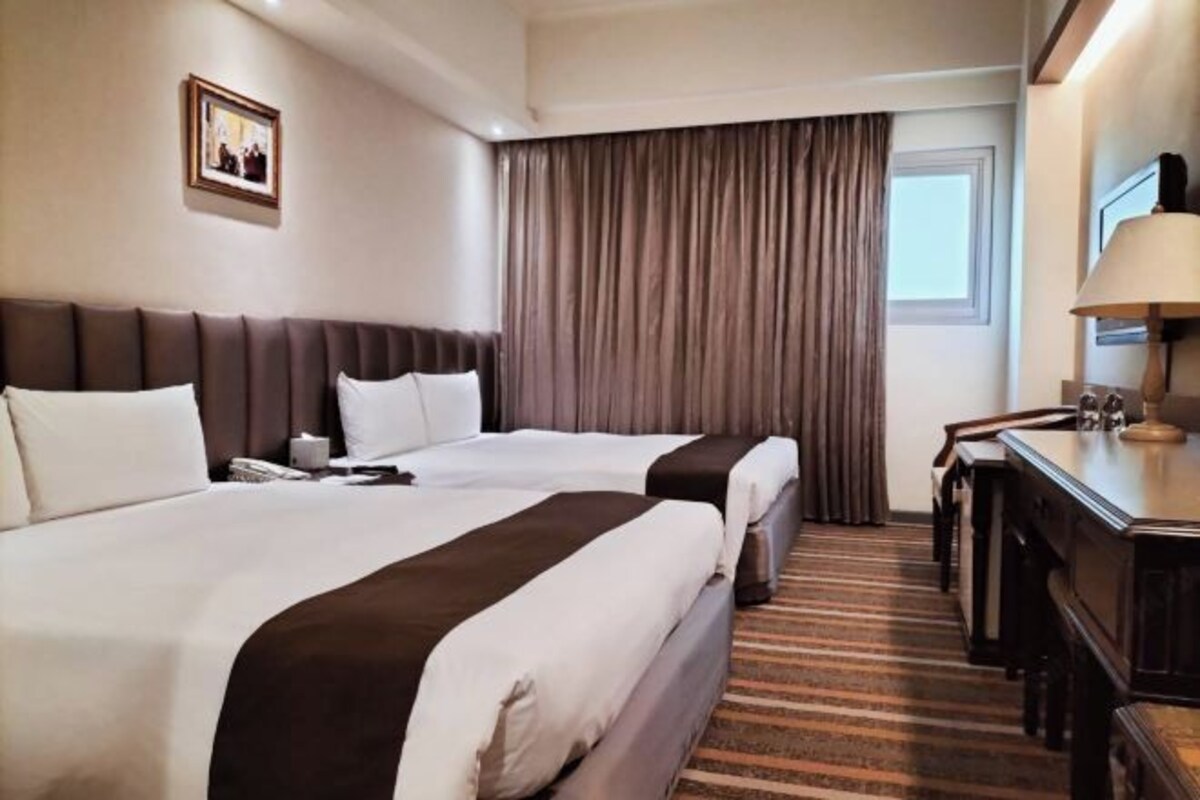 A hotel room with two beds, a desk, a lamp, and a window with curtains.
