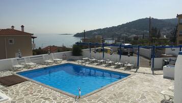 Una piscina con baldosas de piedra, sillones de descanso blancos y vistas al mar y colinas.