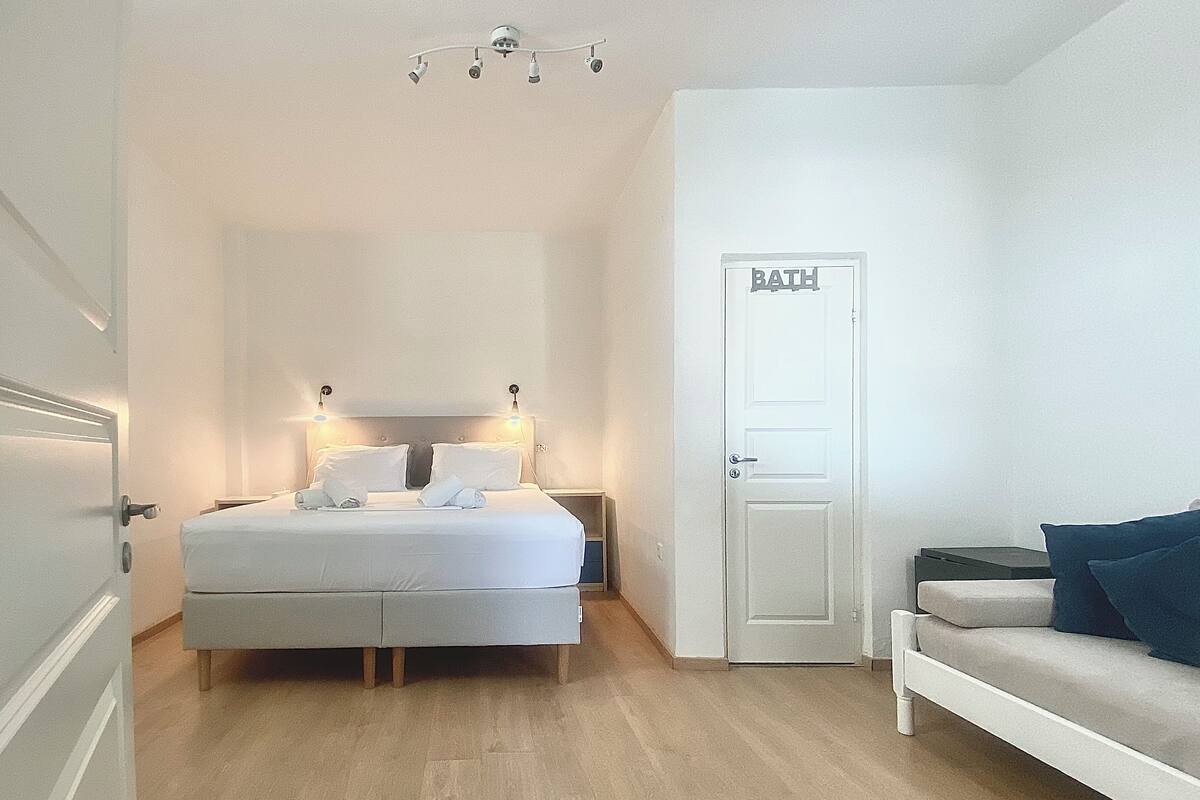 Een hotelkamer met een bed, nachtkastjes, een bank, een deur met daarop 'BATH' en een plafondlamp.
