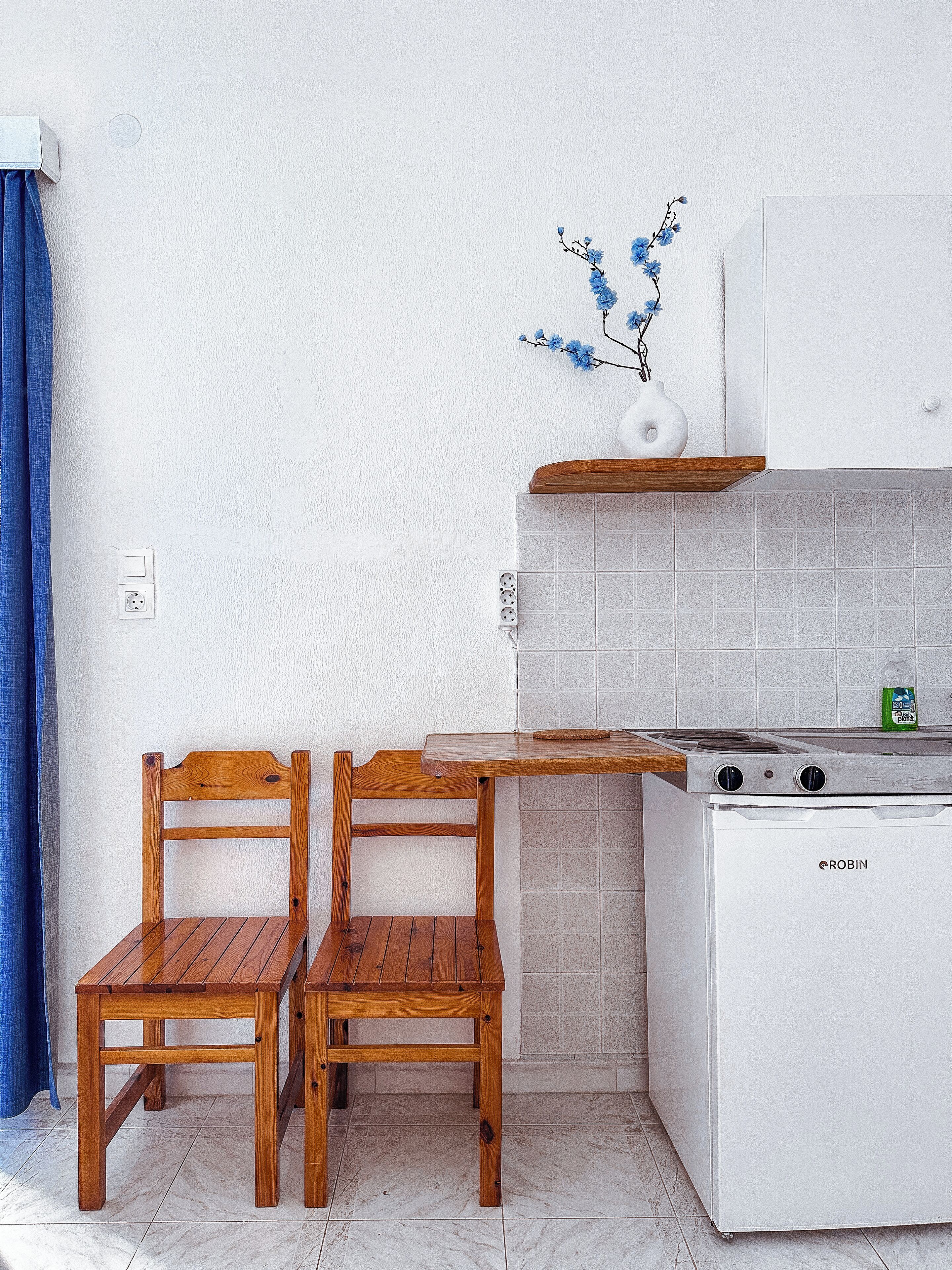 A compact kitchen with a white stove, two wooden chairs, a wooden table, and a blue curtain.