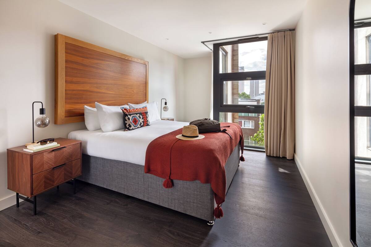 A modern hotel room with a large bed, a nightstand, a window with curtains, and a view of the city.