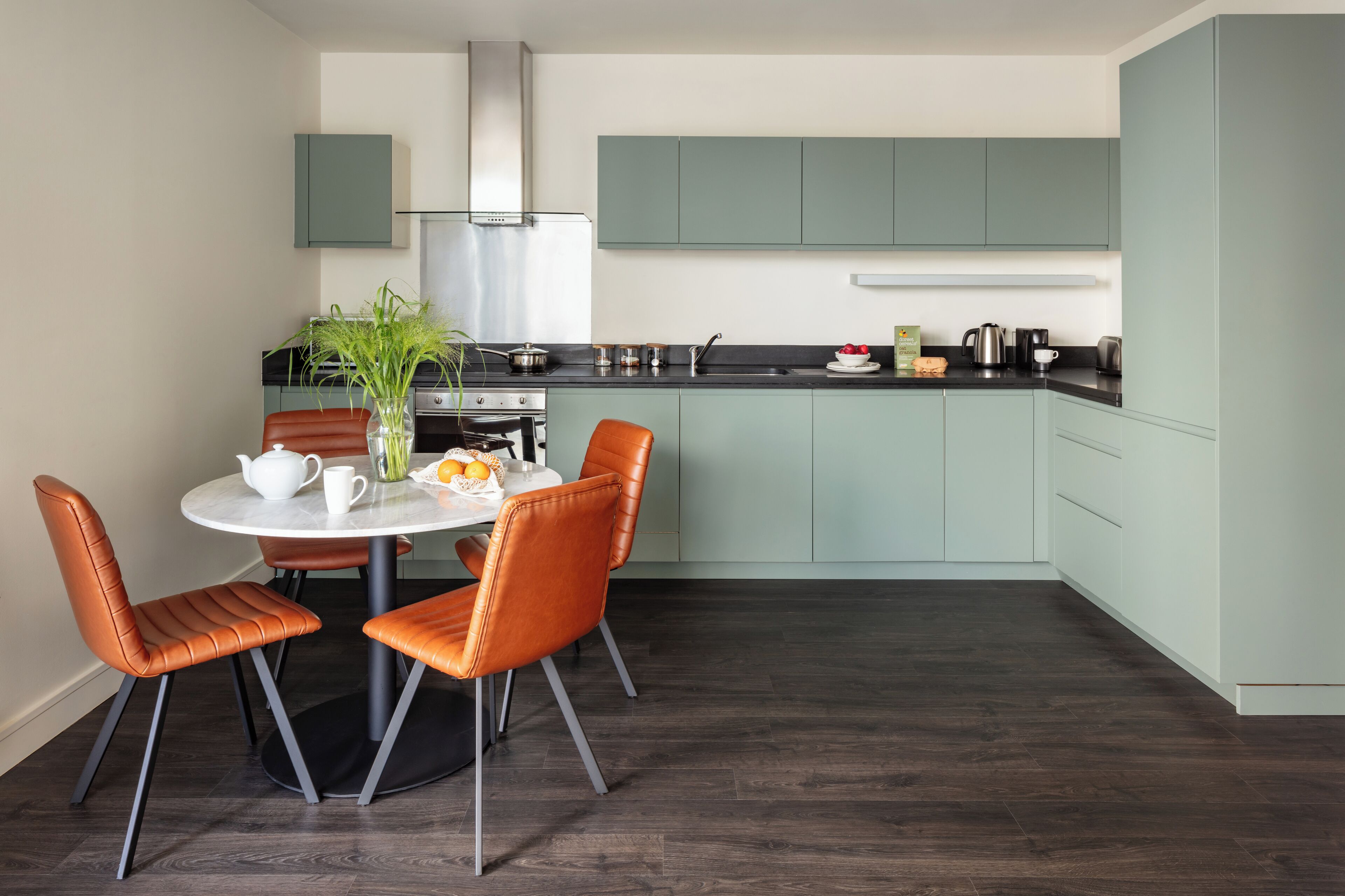 A modern kitchen with teal cabinets, a stainless steel range hood, and a dining area with a round table and orange chairs.