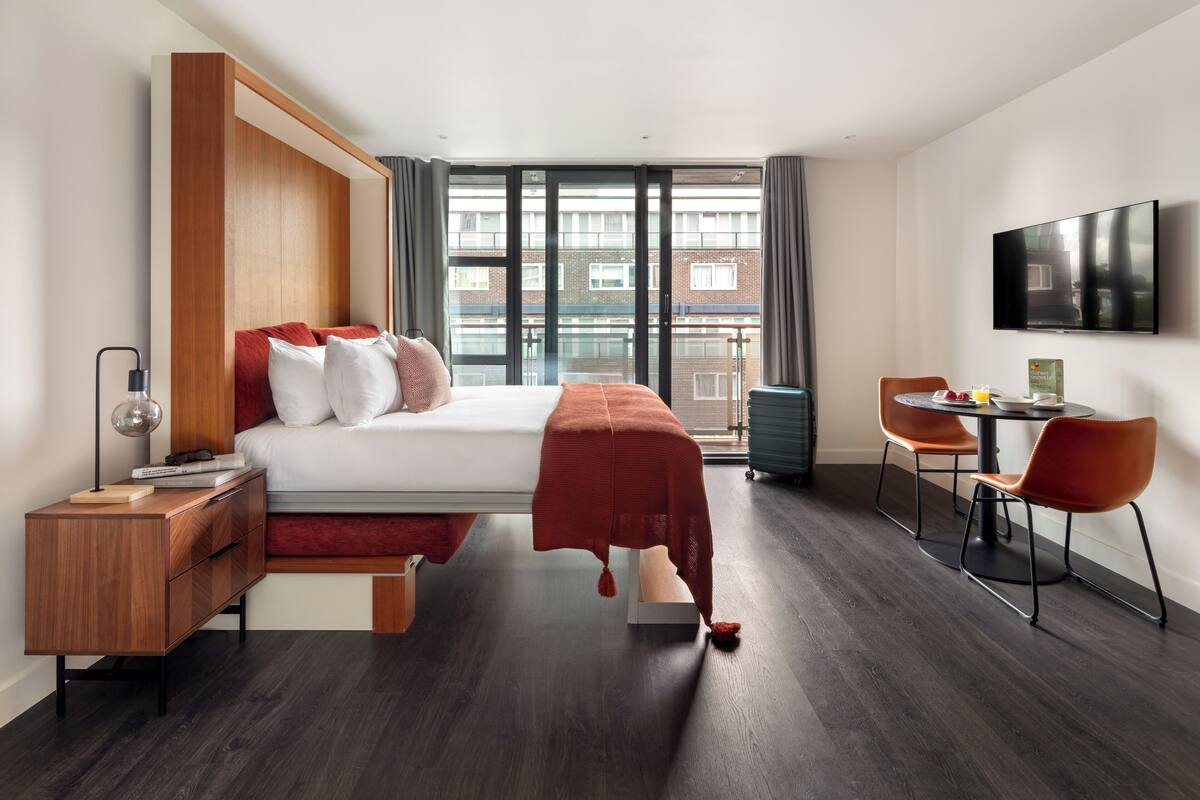 A modern hotel room with a large bed, a desk, a chair, a TV, and a balcony with a city view.