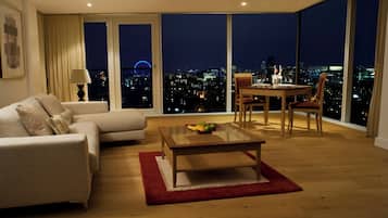 A modern living room with a sofa, a dining table with chairs, a glass coffee table, and a large window with a city view at night.