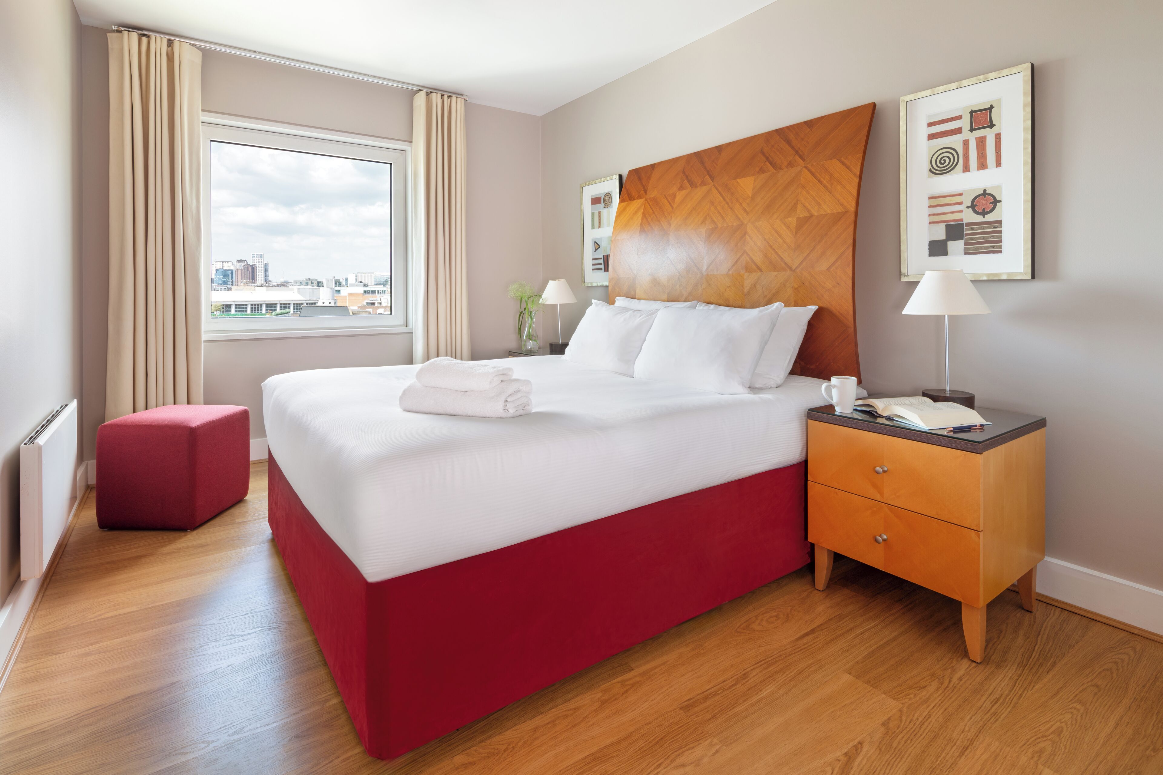 Habitación de hotel con una cama grande, un reposapiés rojo, cabecera de madera, una mesita de noche y vistas a la ciudad desde la ventana.