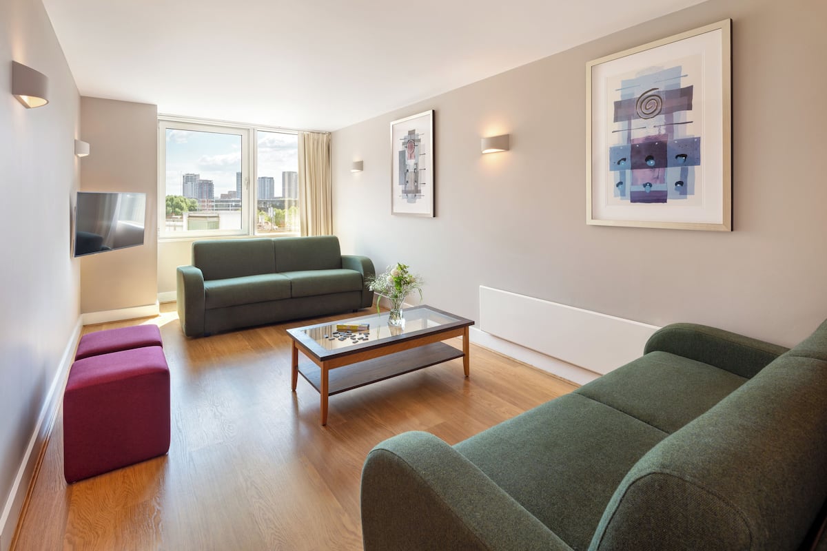 Ein modernes Wohnzimmer mit einem grünen Sofa, einem Glastisch und einem roten Hocker. Ein Fernseher ist an der Wand montiert und über dem Sofa hängt ein abstraktes Bild im Rahmen.
