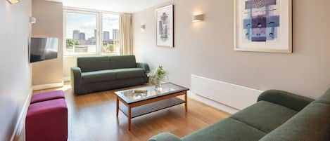 A modern living room with a green sofa, a glass coffee table, and a red ottoman. There is a TV mounted on the wall and a framed abstract painting above the sofa.