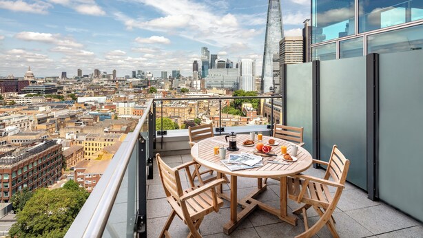 Une terrasse sur le toit, avec une table ronde en bois et des chaises, d’où l’on peut admirer un panorama sur la ville, avec ses bâtiments modernes et son dôme historique.