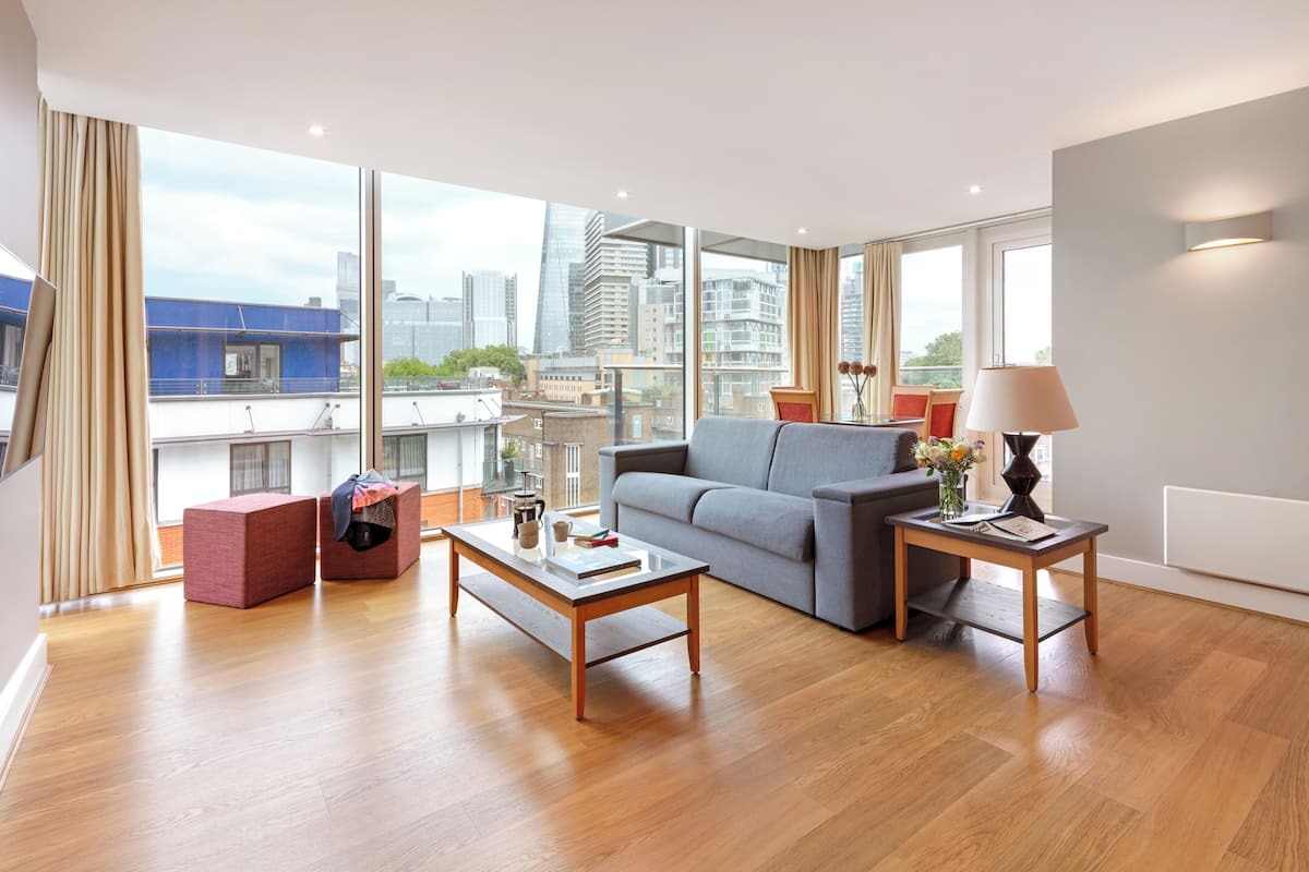 Ein modernes Wohnzimmer mit einem grauen Sofa, einem Holztisch und einem kleinen Beistelltisch mit einer Lampe. Große Fenster bieten Blick auf die Stadtlandschaft.