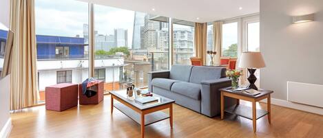 A modern living room with a grey sofa, a wooden coffee table, and a small side table with a lamp. Large windows offer a view of the city skyline.