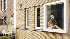 A person sitting by a window with a towel on their head, a brick wall, and outdoor seating with cushions. - Kaboom Maastricht (Maastricht)
