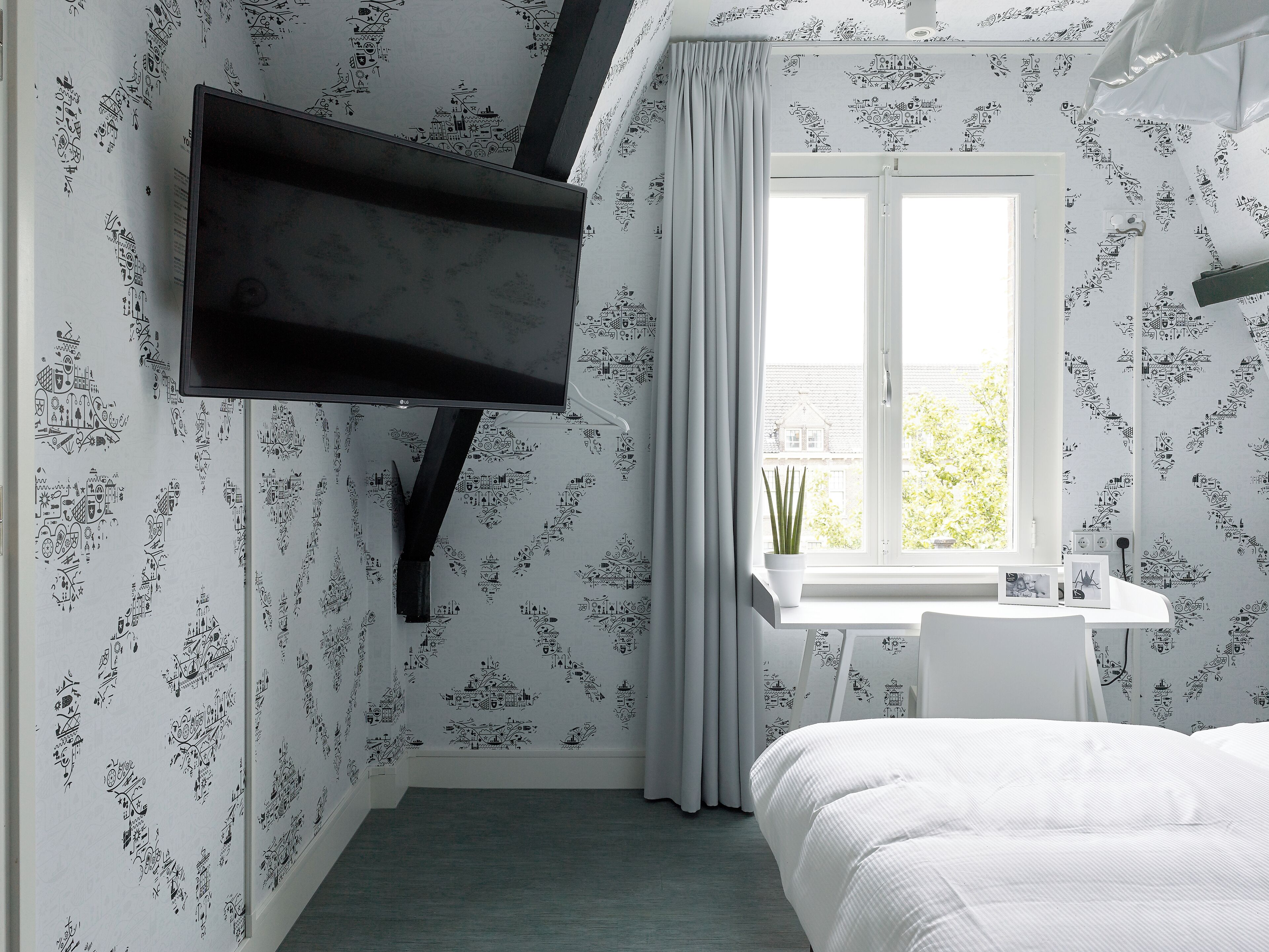 A bedroom with a TV mounted on the wall, a window with curtains, a bedside table, and a bed.