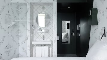 A hotel room with a white bed, a round mirror above the sink, and a dark door.