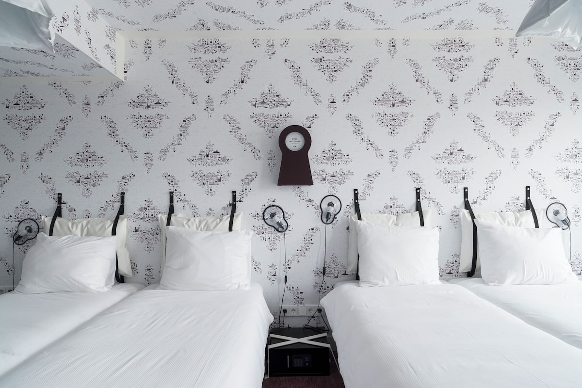 Ein Hotelzimmer mit zwei Betten, weißer Bettwäsche, einem gemusterten Wandteppich und einer an der Wand befestigten Uhr.