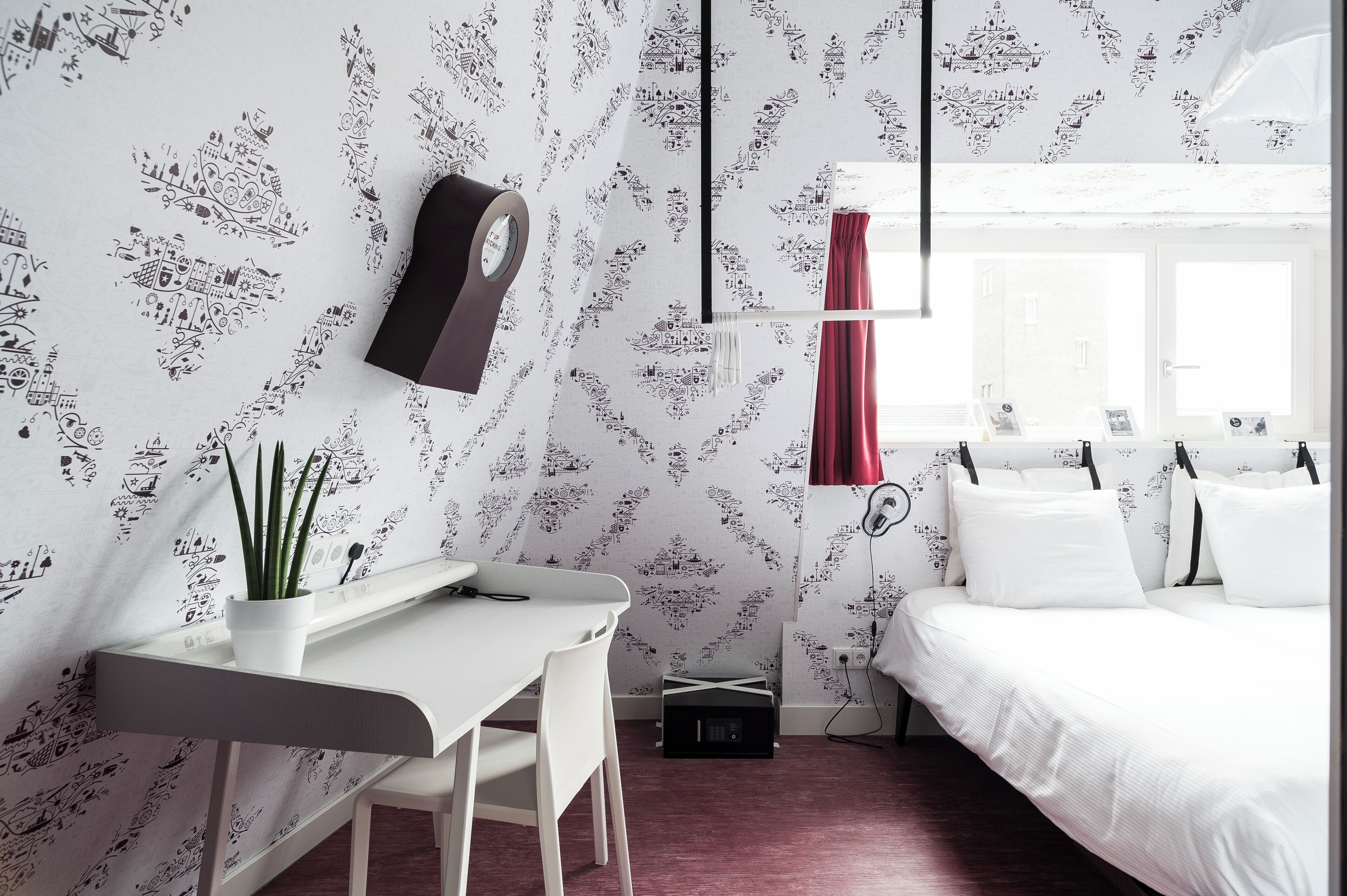 Un dormitorio moderno con una cama blanca, un escritorio y una silla. Hay una cortina roja y un reloj de pared.