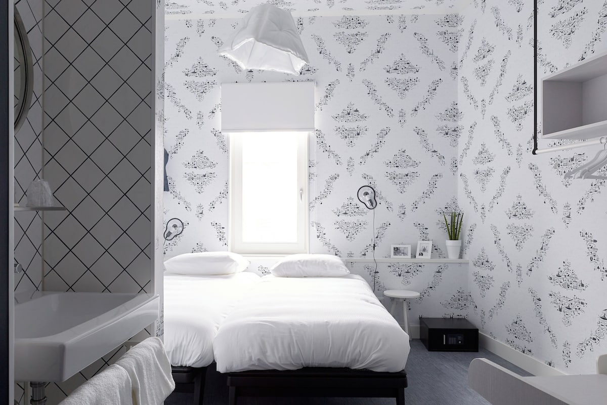 Ein modernes Schlafzimmer mit einem Bett, einem Nachttisch, einem kleinen Fenster und einem linksseitig sichtbaren Badezimmer.