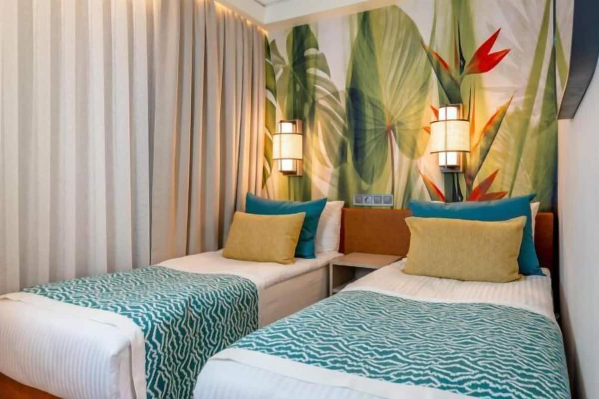A hotel room with two beds, a large tropical-themed wall, and two wall-mounted lamps.