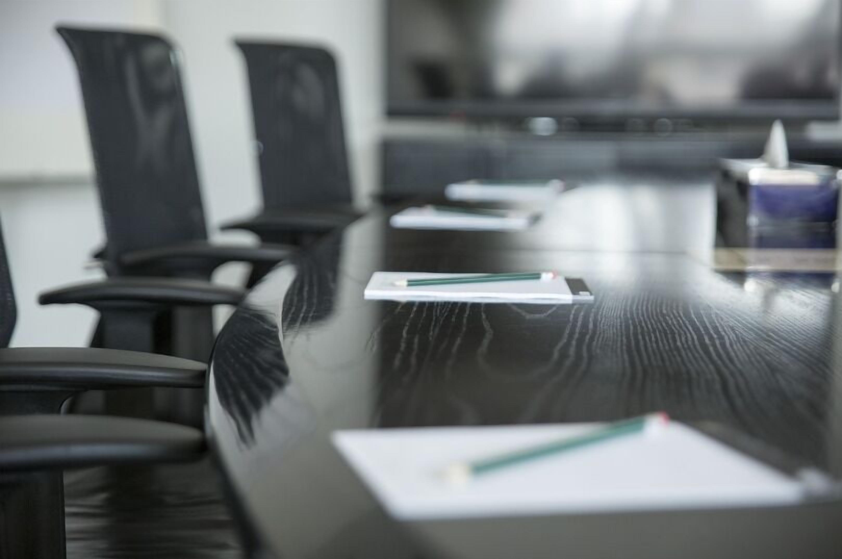 A conference room with a long table, black chairs, and some papers and pens on the table.