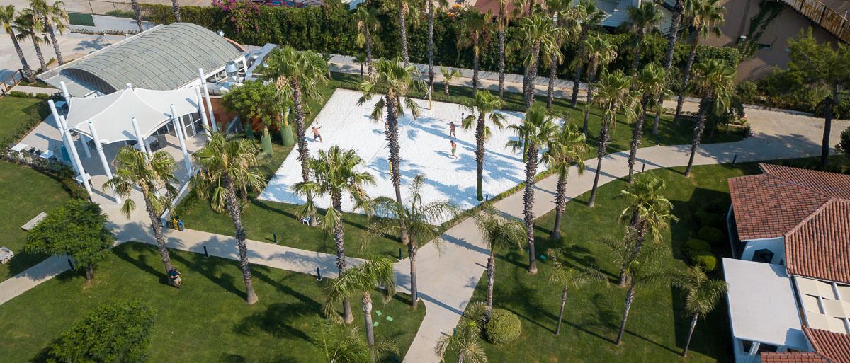 Vista aérea de un resort con una gran piscina rodeada de palmeras, un pabellón cubierto y un edificio con techo de tejas rojas.