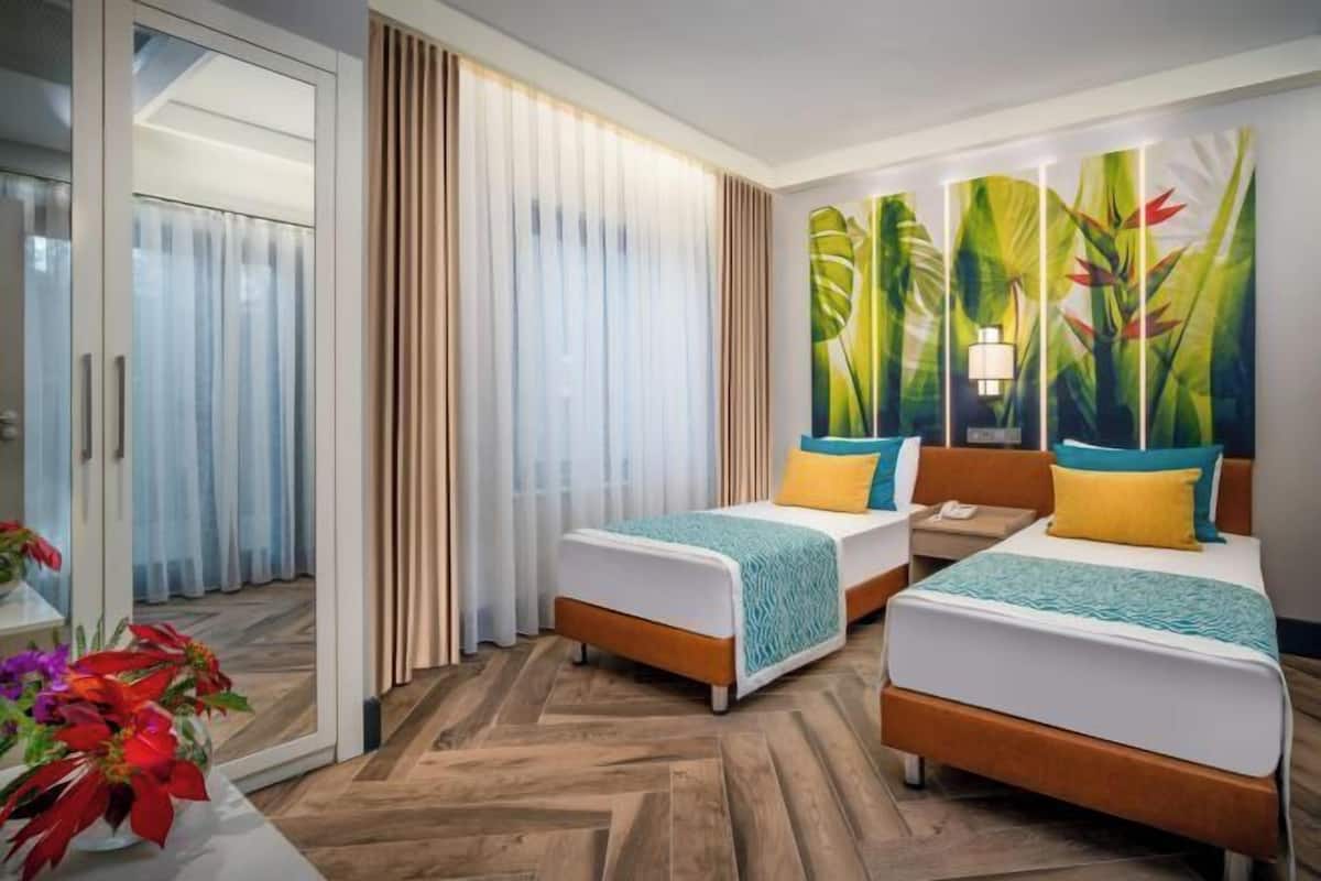 A hotel room with two beds, a sliding glass door, a large tropical-themed painting, and a wooden herringbone floor.