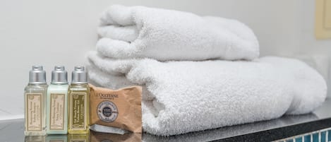 A bathroom with a stack of white towels, a bottle of shampoo, a bottle of conditioner, and a bottle of body lotion on the counter.