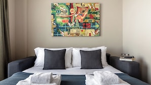A hotel room with a bed, two pillows, a bedside table with books, and a colorful abstract painting on the wall.