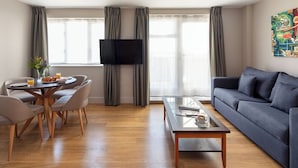 A modern living room with a sofa, a coffee table, a dining table with chairs, a flat-screen TV mounted on the wall, and large windows with curtains.