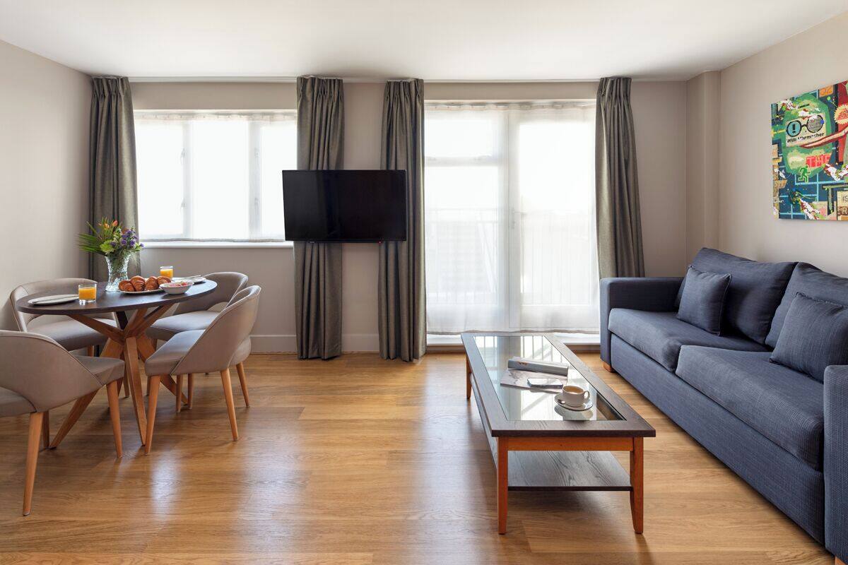 Ein modernes Wohnzimmer mit einer Couch, einem Couchtisch, einem Esstisch mit Stühlen, einem flachbildfernseher an der Wand und großen Fenstern mit Vorhängen.