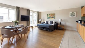 A modern hotel room with a bed, a dining area with chairs and a table, a kitchen area, and a large window with curtains.