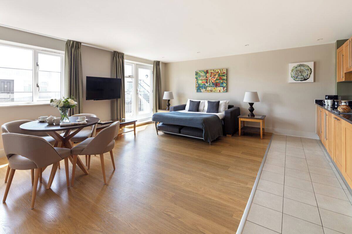 Ein modernes Hotelzimmer mit Bett, einer Essecke mit Stühlen und Tisch, einer Küchenzeile und einem großen Fenster mit Vorhängen.