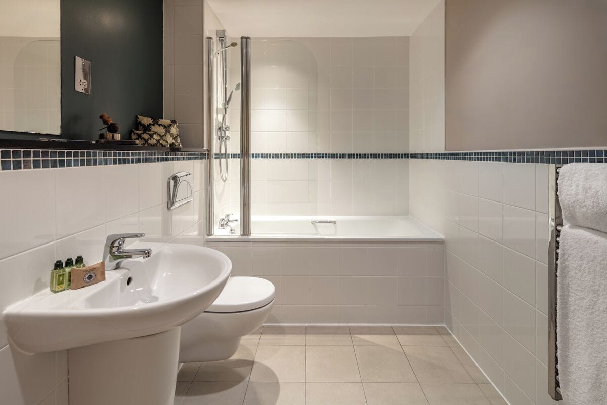 A modern bathroom with a white sink, toilet, and bathtub. There is a shower area with a glass door. The walls are tiled, and there is a towel rack on the right.