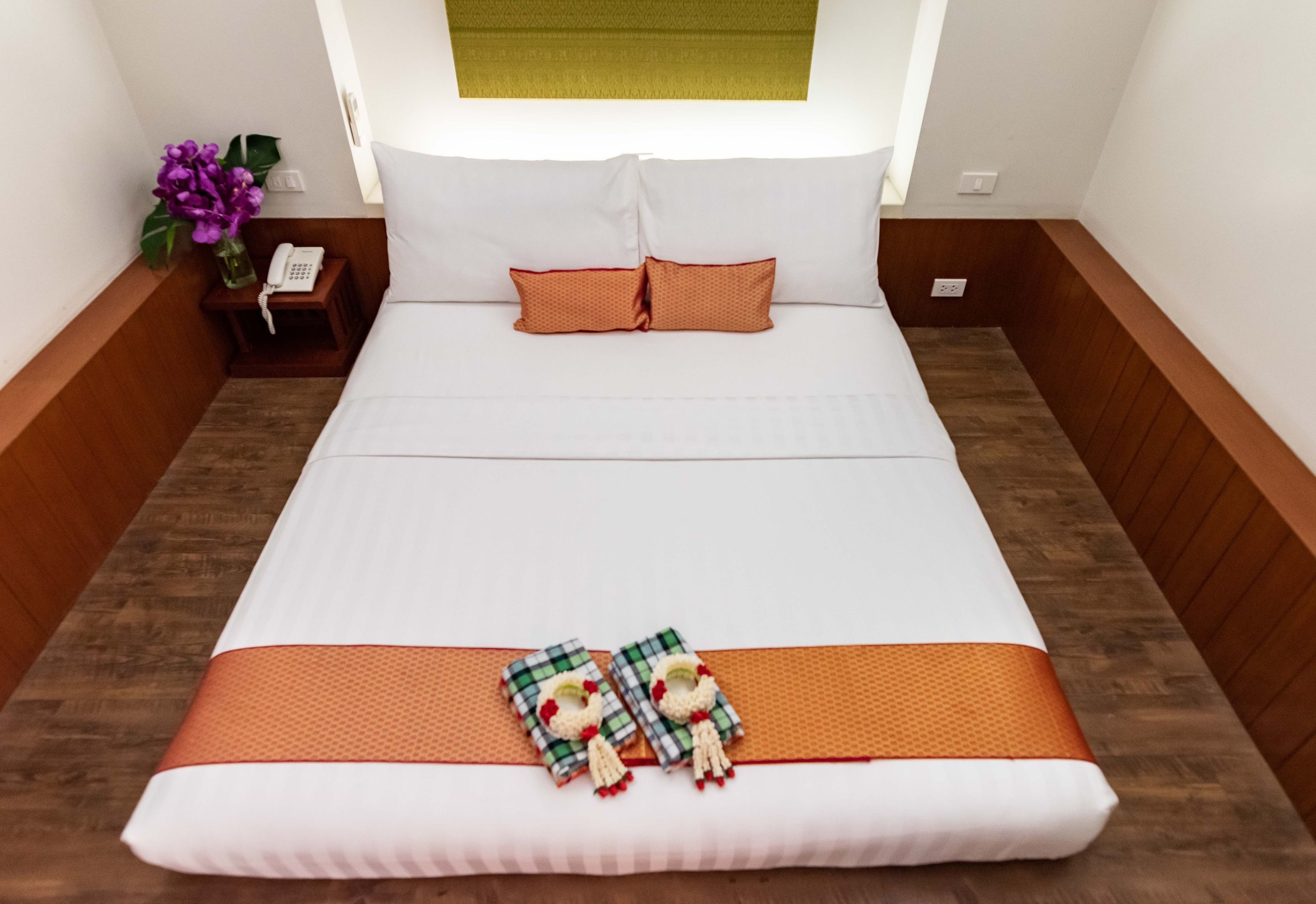 A neatly made bed with a white comforter, two orange pillows, and a decorative blanket with a checkered pattern.