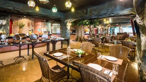 Um restaurante com mesas altas e cadeiras de vime, parede de tijolos com uma planta e luzes decorativas penduradas.
