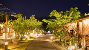 A pathway flanked by potted plants and illuminated by soft lighting, leading to a building with a covered entrance.