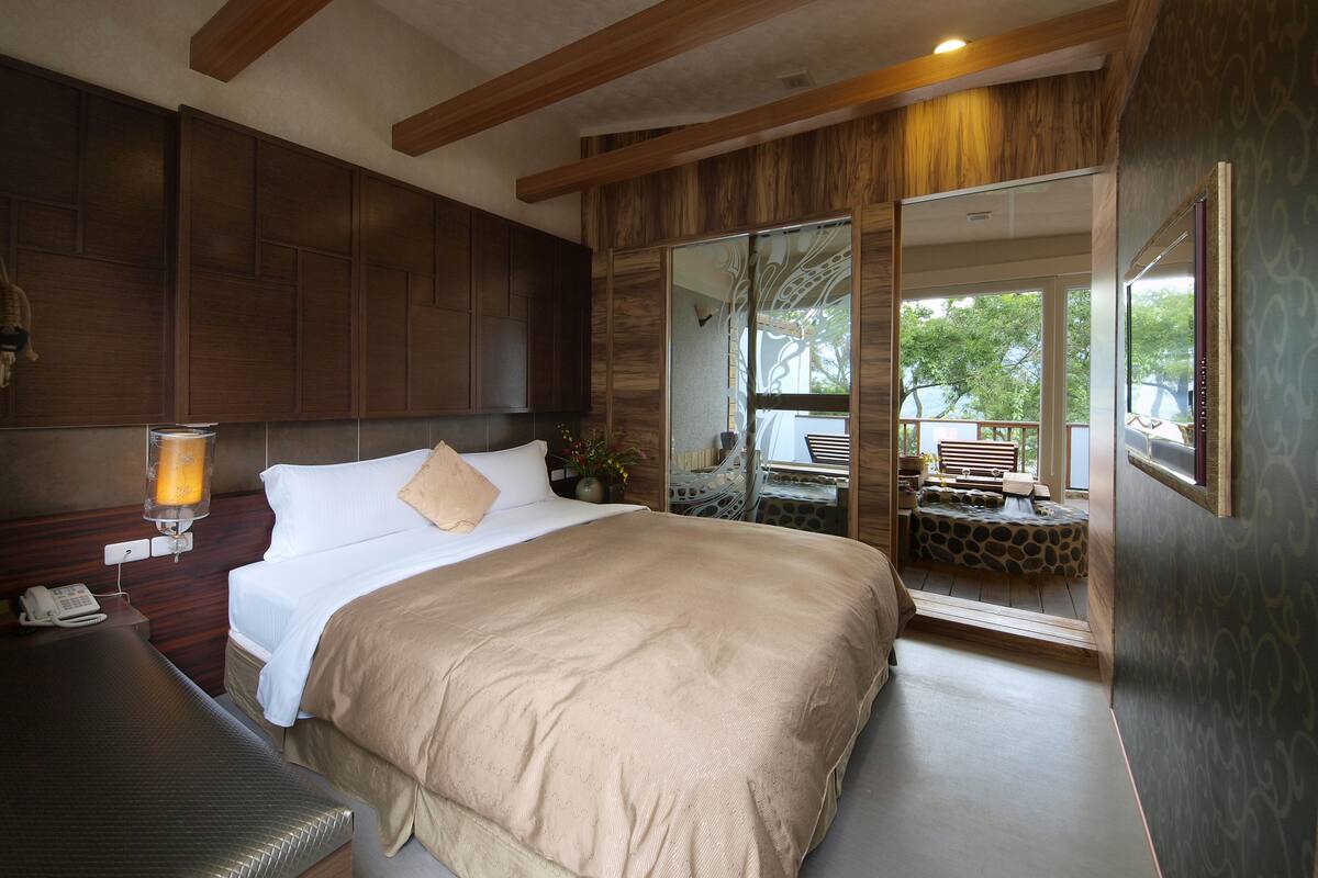 Une chambre d’hôtel avec un grand lit, des poutres apparentes en bois au plafond, un téléphone sur la table de chevet et une vue sur le balcon extérieur.