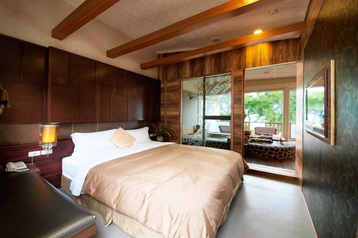 A bedroom with a large bed, wooden headboard, bedside table with a lamp, and a view of the outdoors through a sliding door.