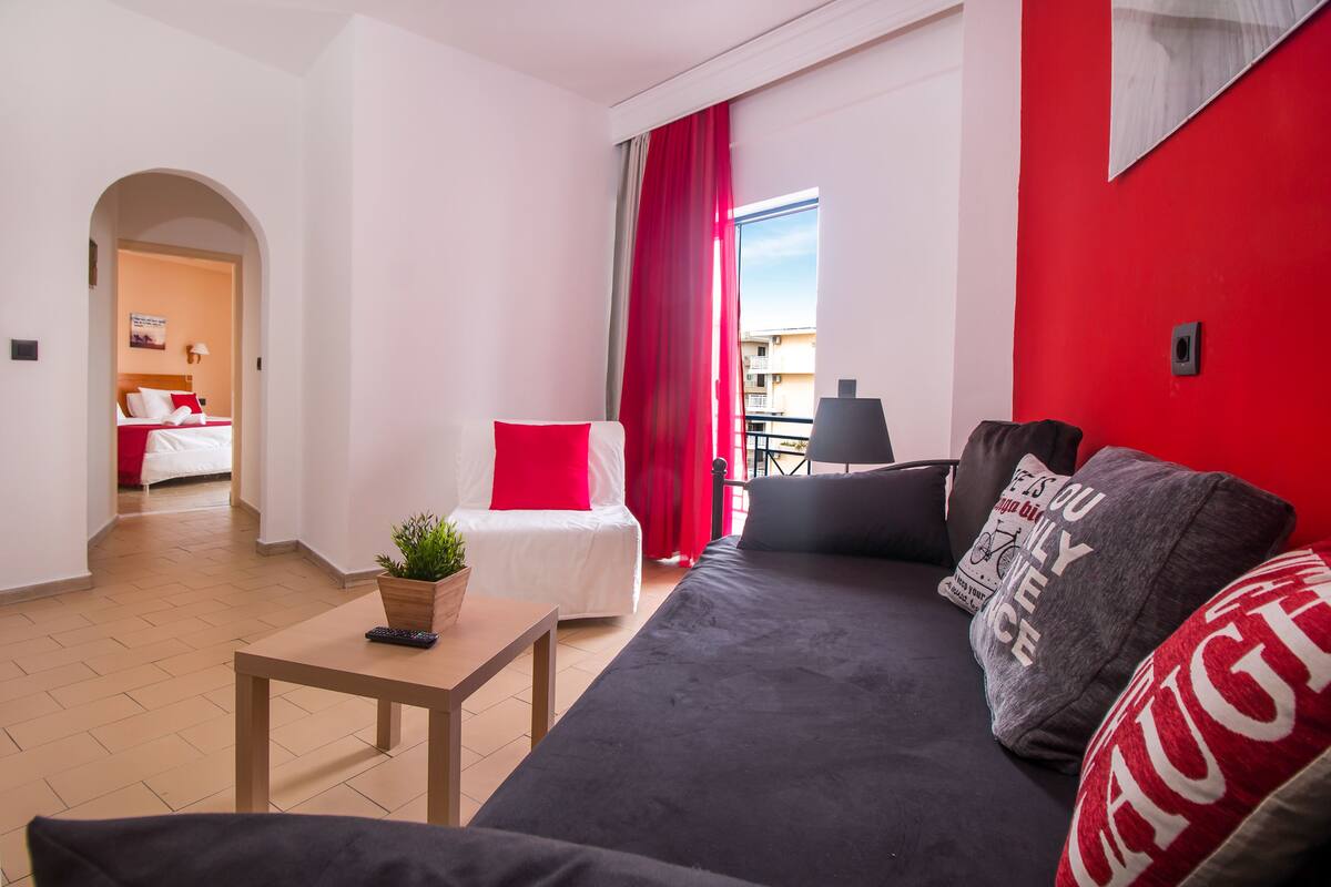 Una sala moderna con un sofá, una mesita con una planta y una cama con ropa de cama roja.