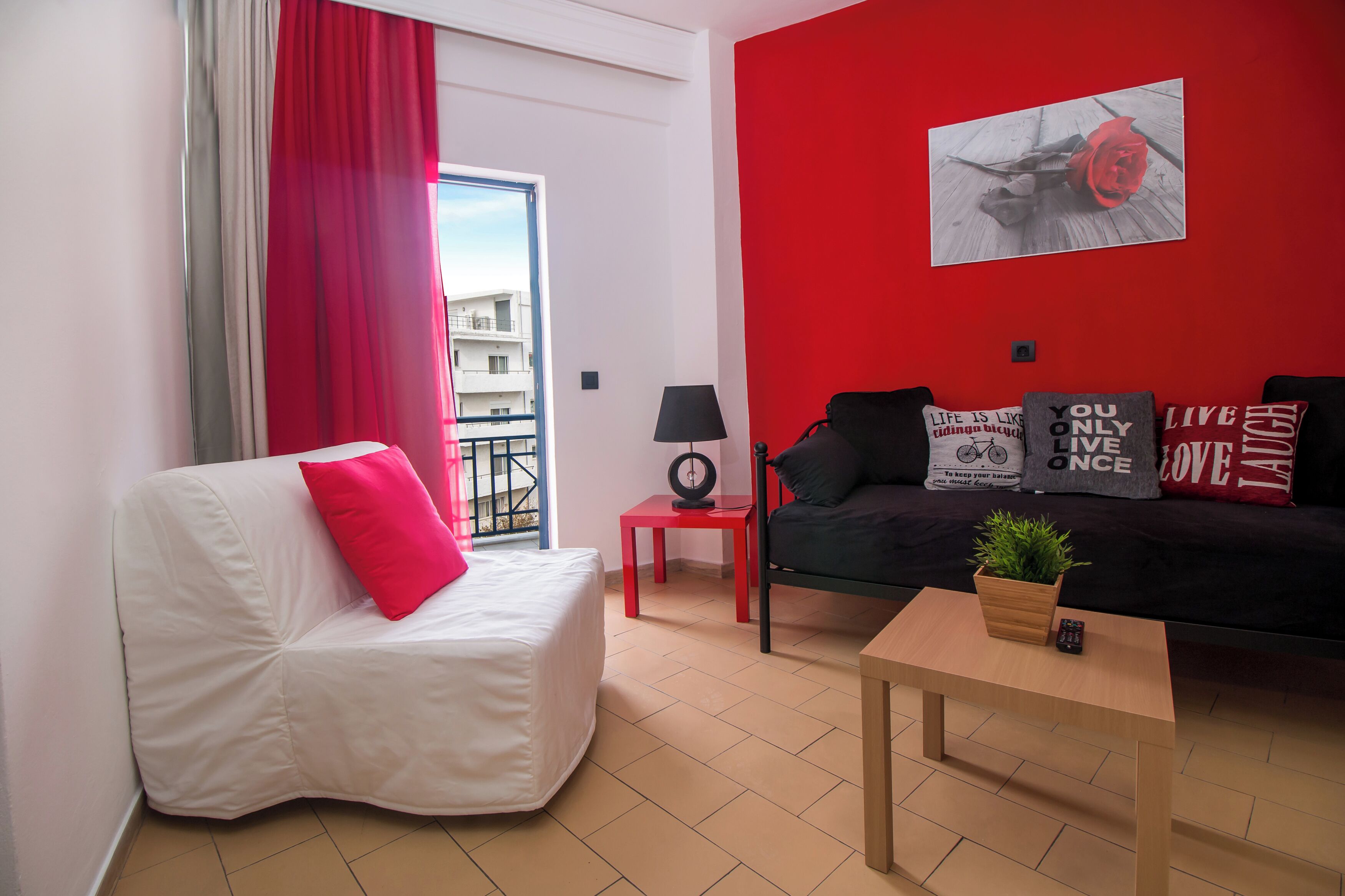 A modern living room with a red accent wall, a white sofa, a black sofa, a small table with a plant, and a red side table with a lamp.
