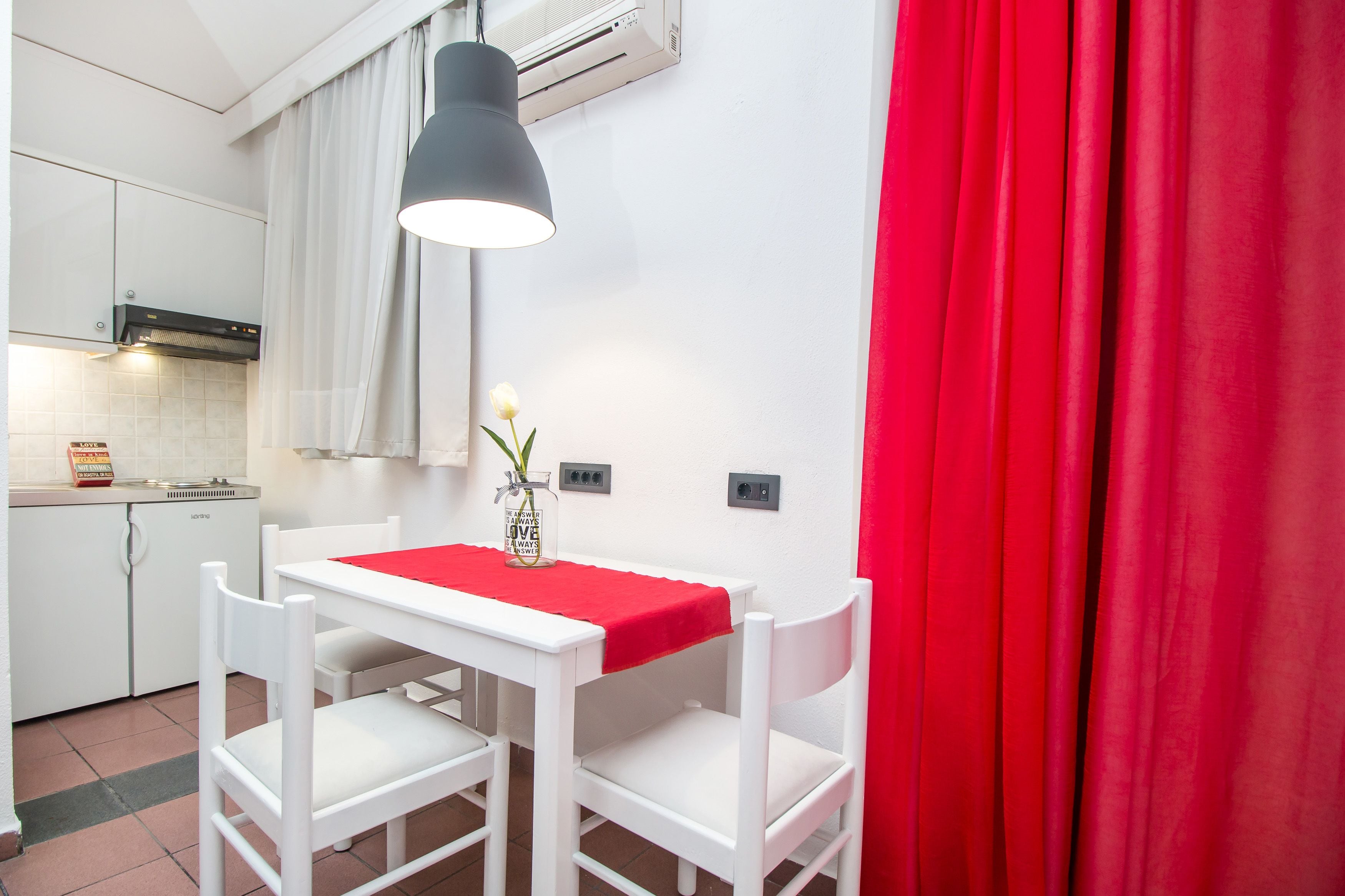 A small dining area with a table and two chairs, a red tablecloth, a vase with a flower, and a black pendant light.