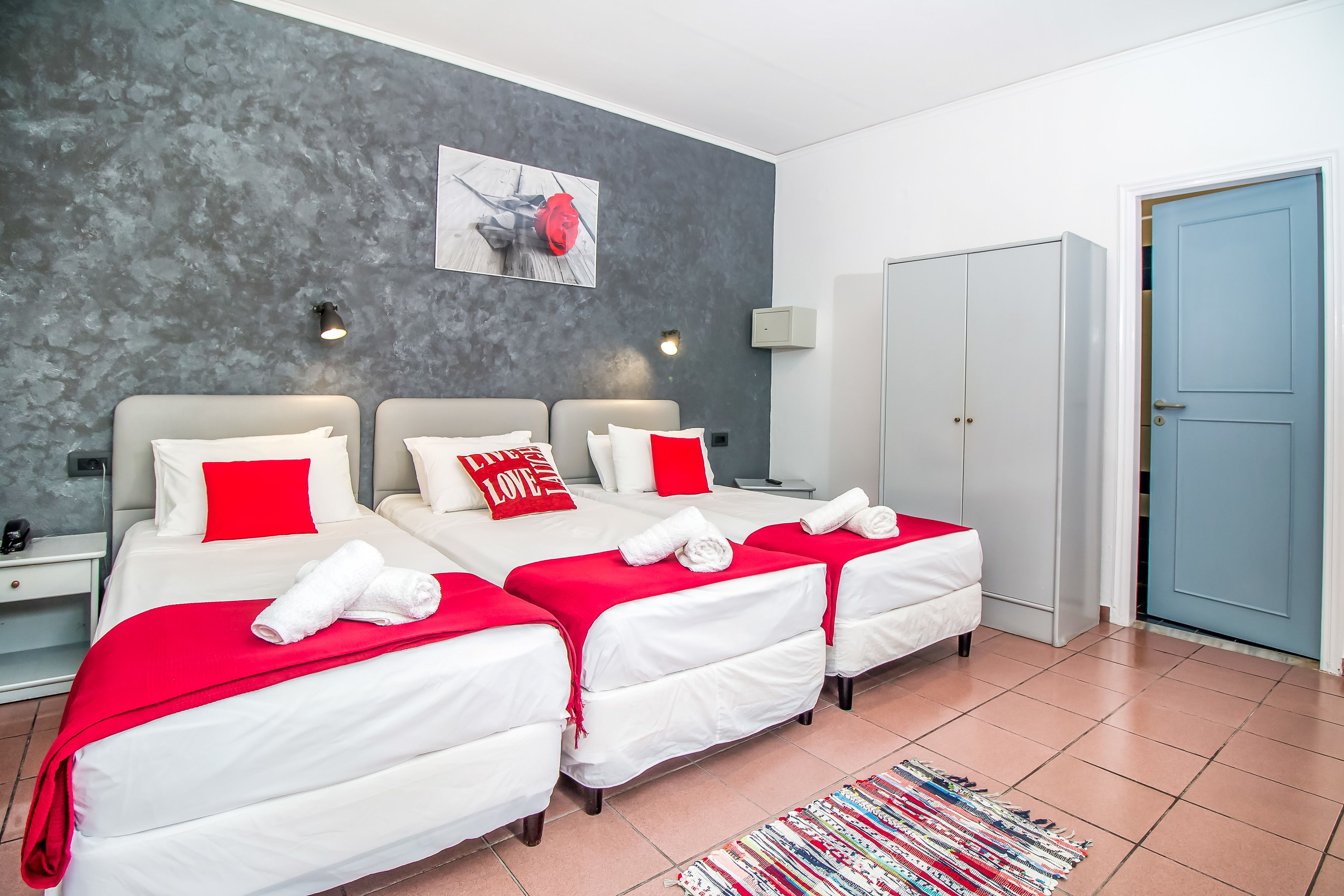 A hotel room with three beds, each with white bedding and red pillows, a grey textured wall, a painting, and a blue door.