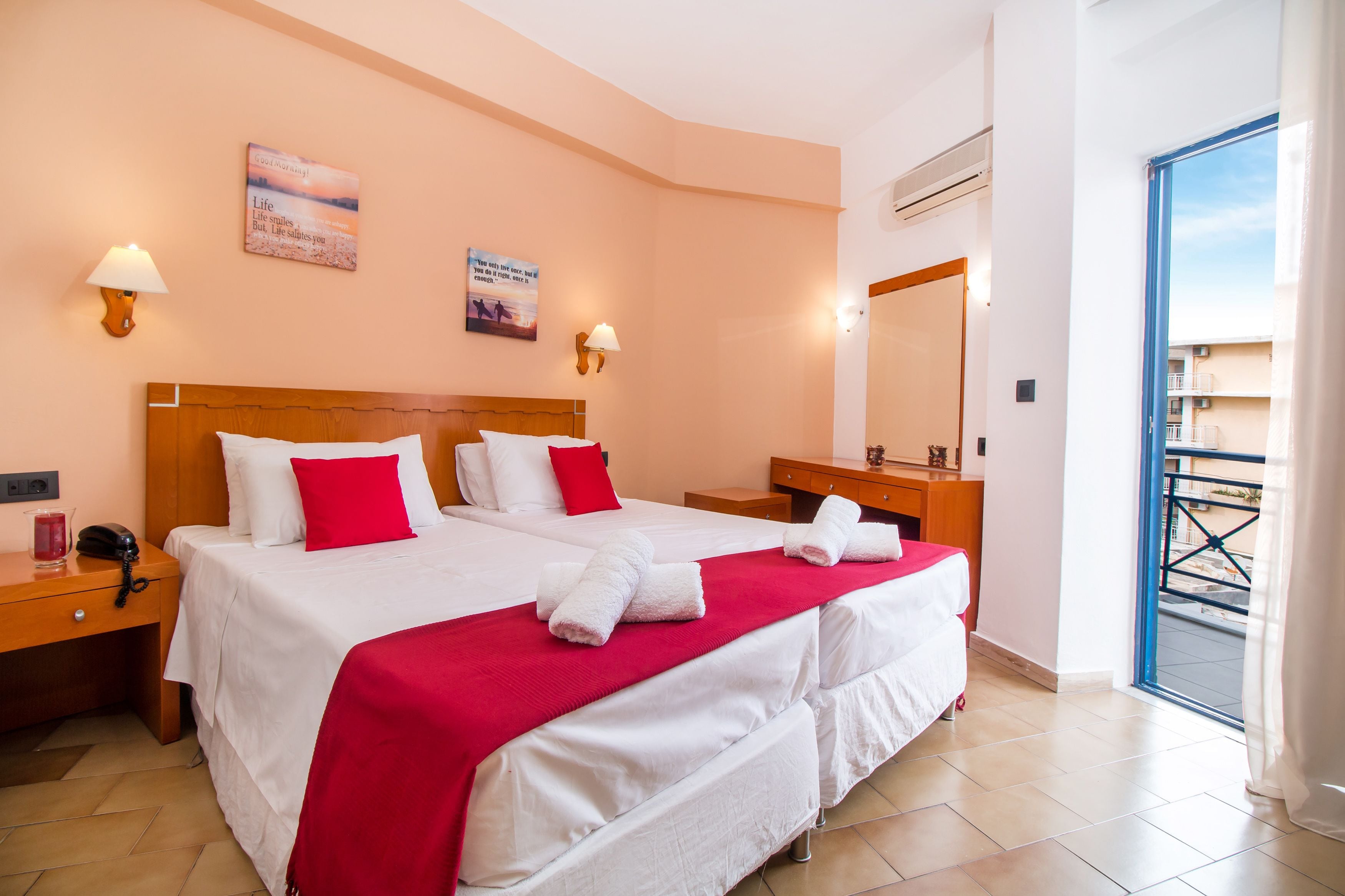 A hotel room with two beds, a wooden headboard, a red throw blanket, and a balcony with a view.