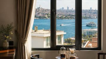 Quarto de hotel com uma cama grande, uma mesa de centro com cafeteira e chávenas, vista para a paisagem urbana e para a água através de uma janela.