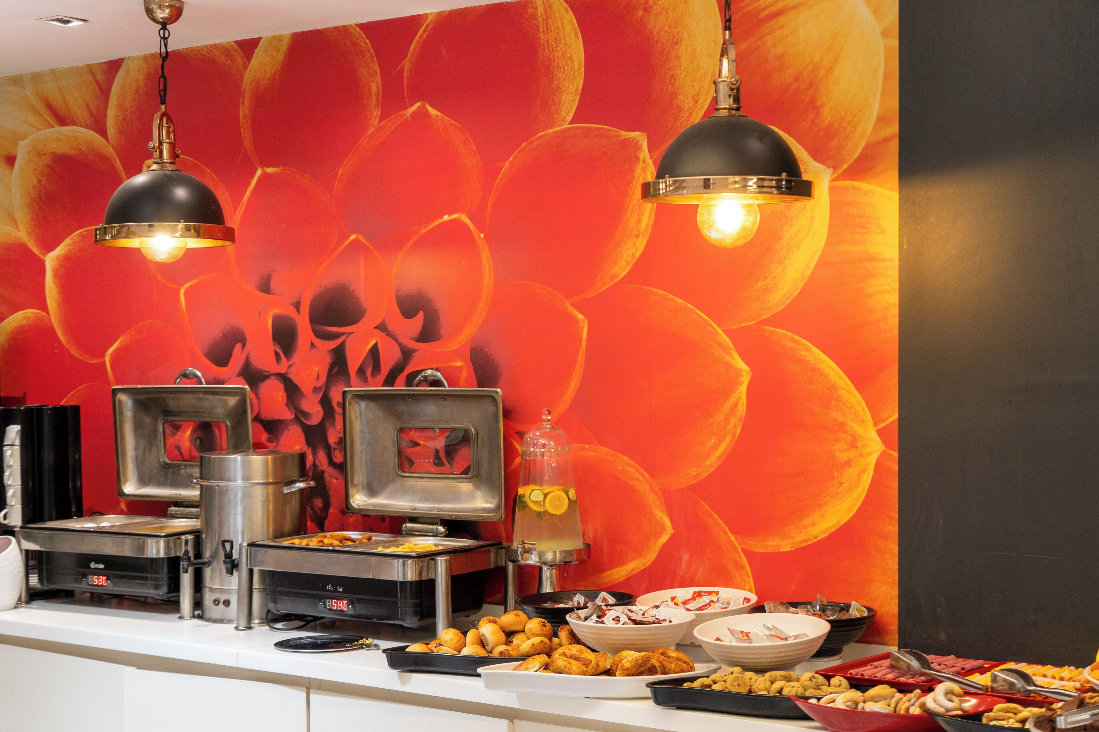 Disposição de buffet com diversas estações de comida, uma máquina de café e uma parede decorativa com padrão floral.