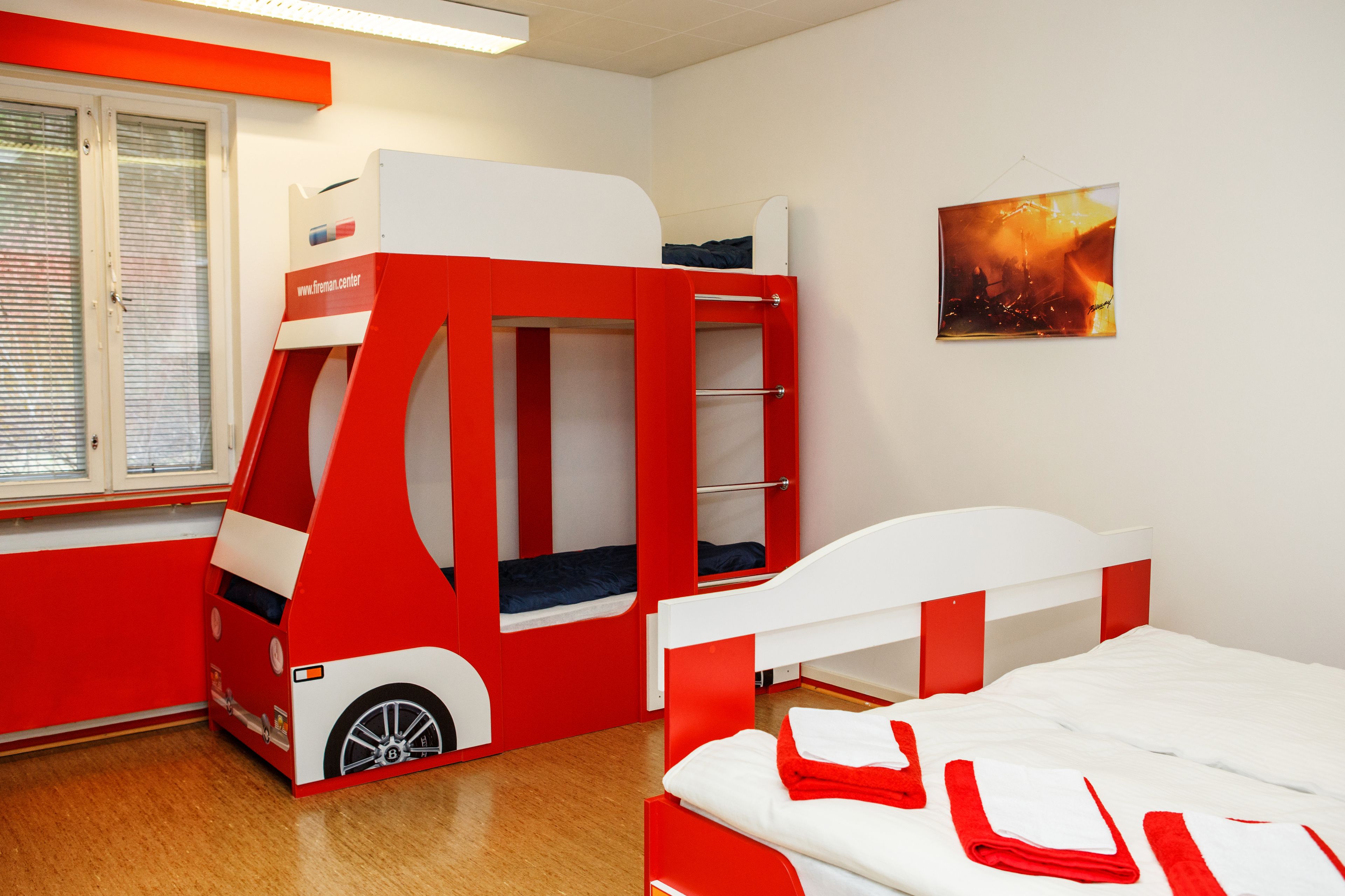 Une pièce comprenant un lit superposé, un petit lit superposé fermé, et un lit simple. Il y a une fenêtre avec des stores, une porte et un tableau accroché au mur.