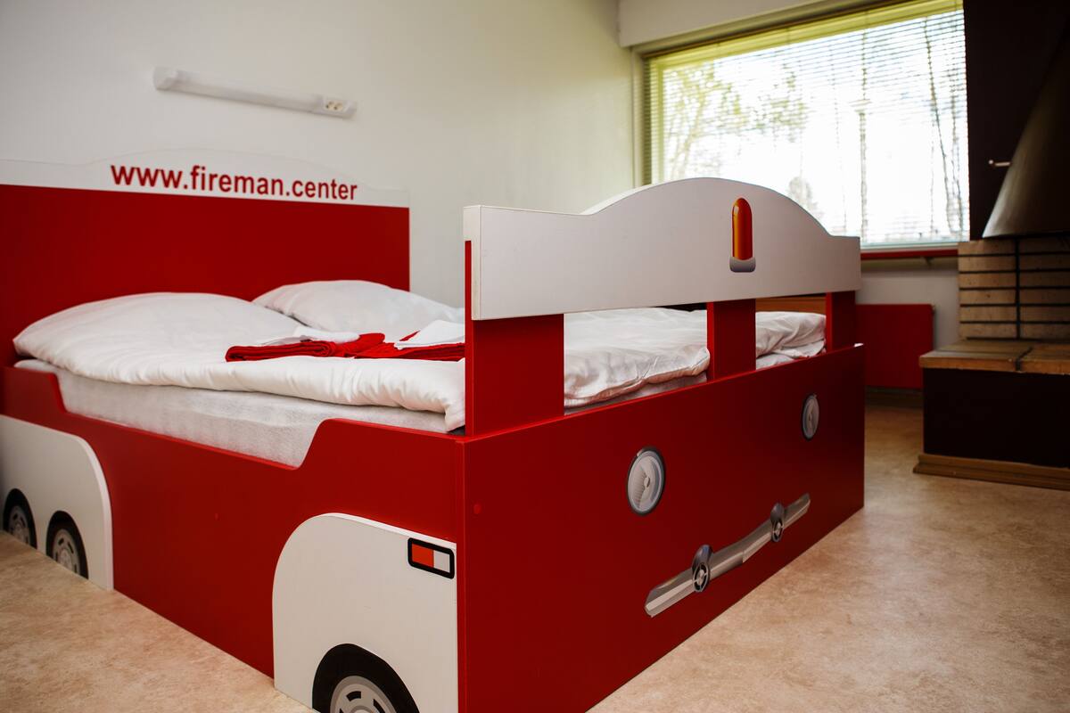 A red and white bunk bed with a headboard resembling a fire truck, a window with blinds, and a website address on the wall.