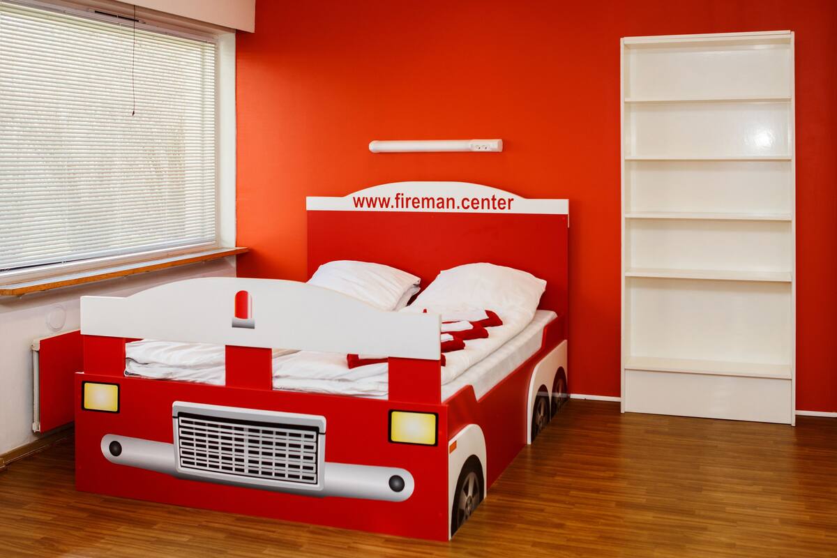 A room with a red and white bed designed like a fire truck, a window with blinds, and a white shelf unit against an orange wall.