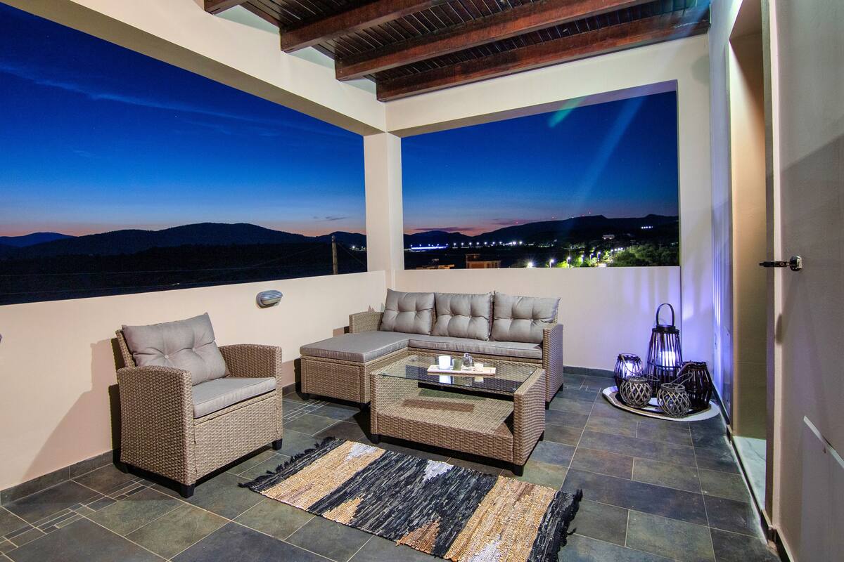 A balcony with wicker furniture, a glass table, and a view of the mountains at dusk.