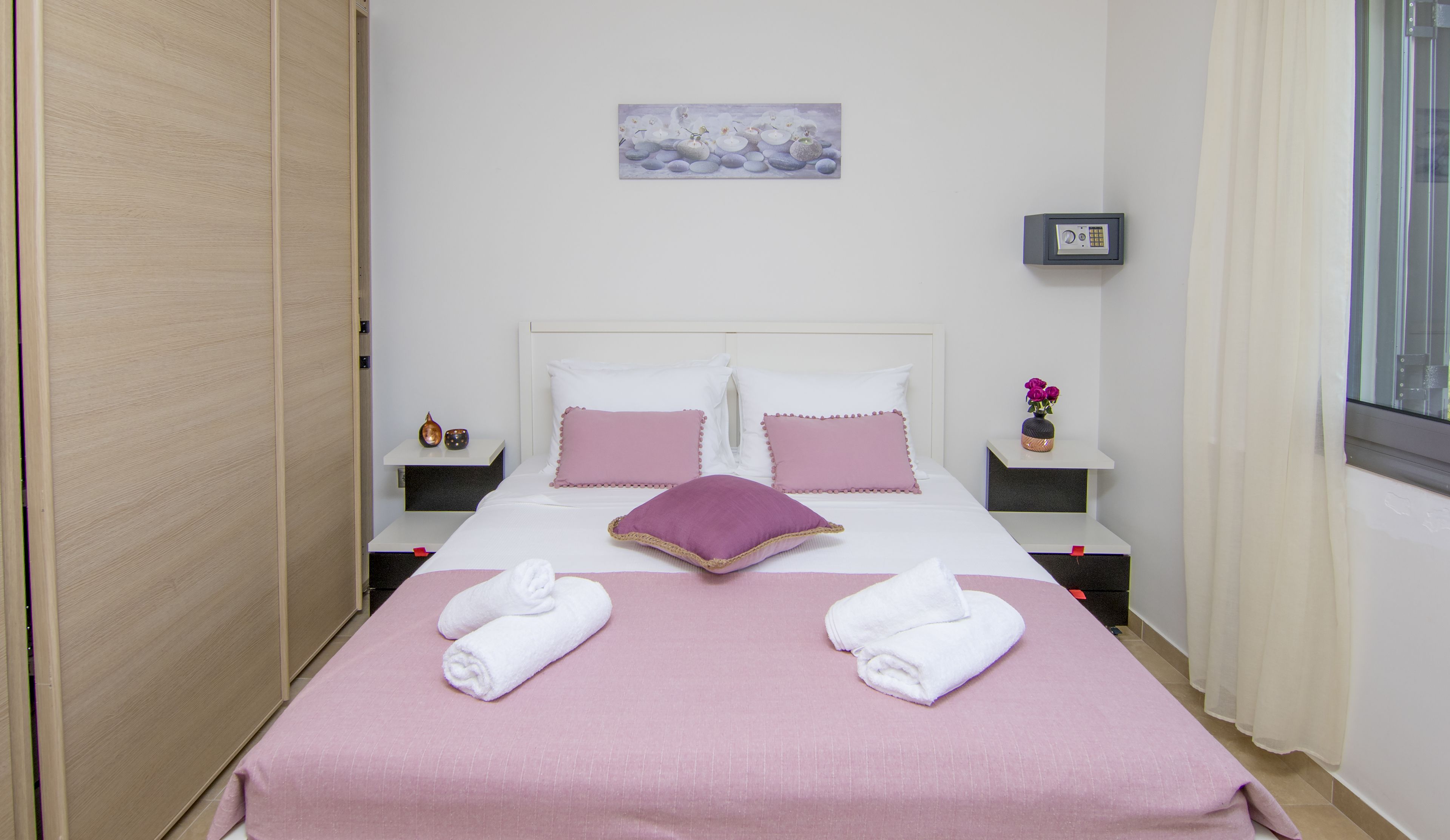 A neatly arranged bedroom with a bed, pillows, a bedside table, a nightstand, a painting, a safe, and towels.