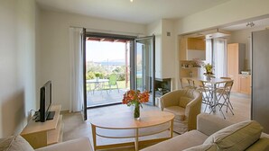 A modern living room with a sofa, a coffee table, and a TV. There is a sliding glass door leading to a balcony with a view of the outdoors. - Haraki Villas (Rhodes)