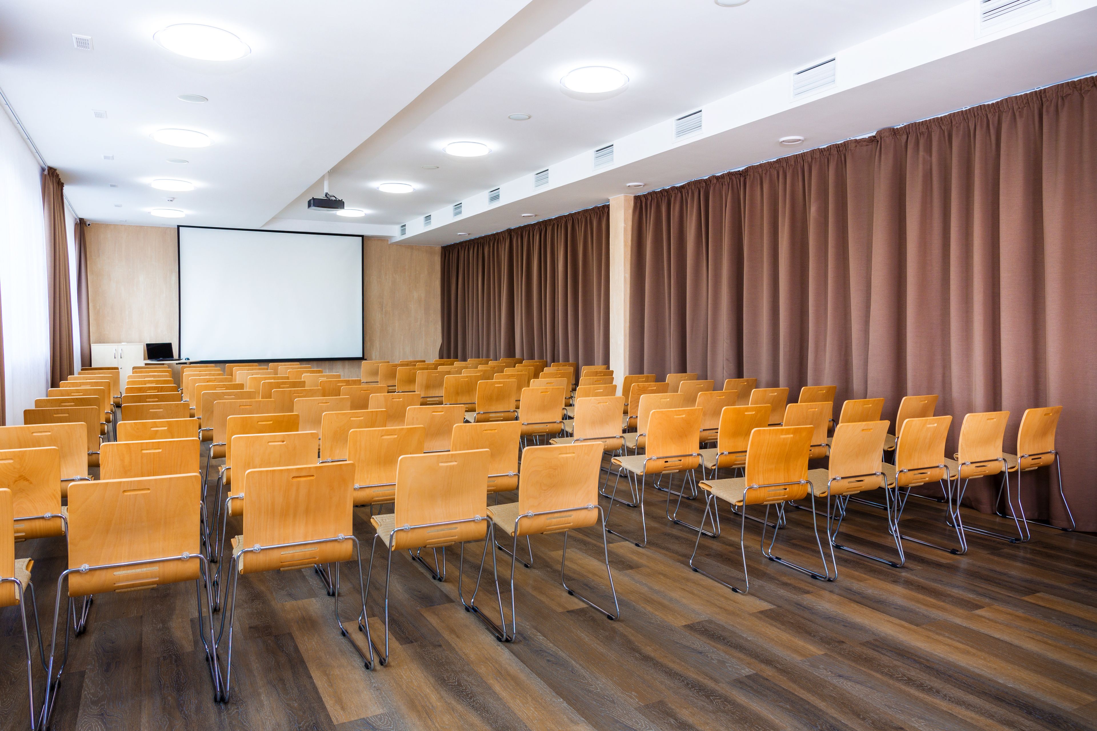 Une salle de conférence avec des rangées de chaises, un écran de projection et un plancher en bois.